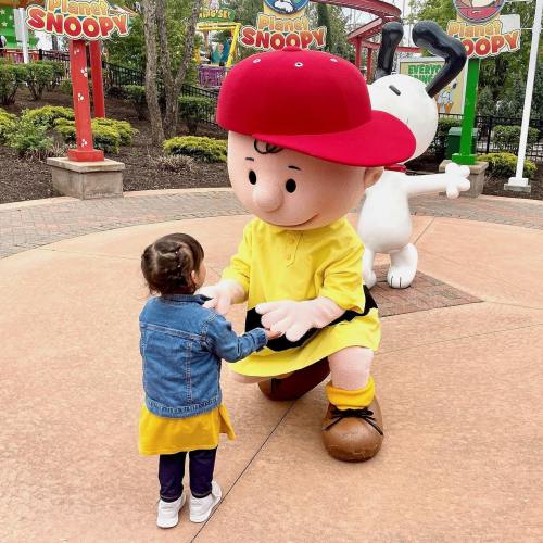 A young child in a yellow dress and denim jacket interacts with a large cartoon character in a red hat at a theme park.