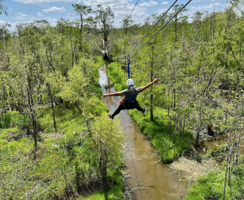 A person zip-lining over a lush green forest with a stream below, arms outstretched and wearing a helmet.