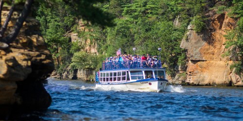 A tour boat filled with passengers navigates a river surrounded by lush greenery and rocky cliffs.