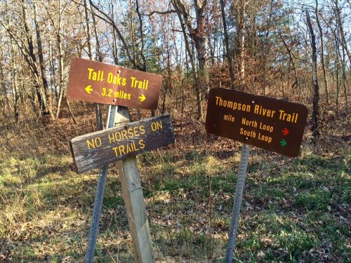 Two wooden trail signs in a wooded area, one for Tall Oaks Trail and the other for Thompson River Trail, with a "No Horses" sign.