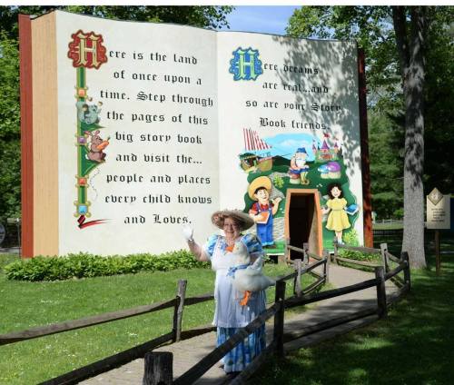 A woman in a hat stands in front of a large storybook mural in a park, inviting visitors to explore its pages.