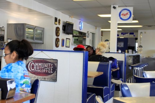 A cozy diner interior with blue and white decor, featuring patrons seated at tables and a sign for community coffee.
