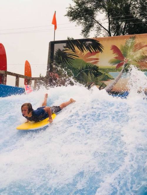 A child rides a yellow boogie board on a wave at a water park, surrounded by colorful surfboards and tropical decor.