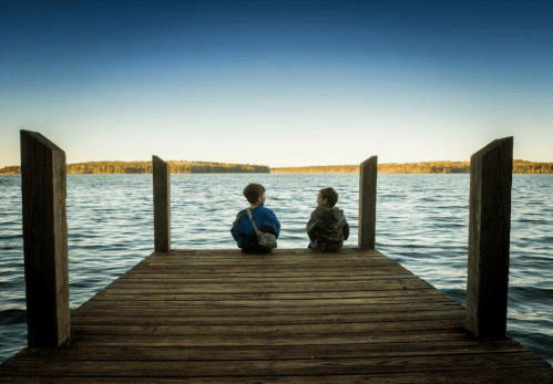 Two children sit on a wooden dock, gazing out at a calm lake surrounded by trees under a clear blue sky.