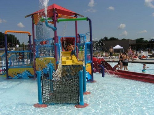 Colorful water play structure with slides and a splash area, surrounded by people enjoying a sunny day at the pool.
