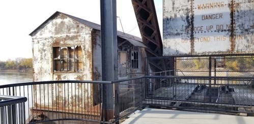 Rusty, abandoned building near a river, with a warning sign and a metal railing in the foreground.