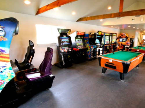 A game room featuring arcade machines, a pool table, and bright lighting with wooden beams on the ceiling.