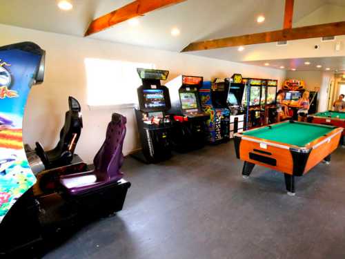 A game room featuring arcade machines, a pool table, and bright lighting with wooden beams on the ceiling.