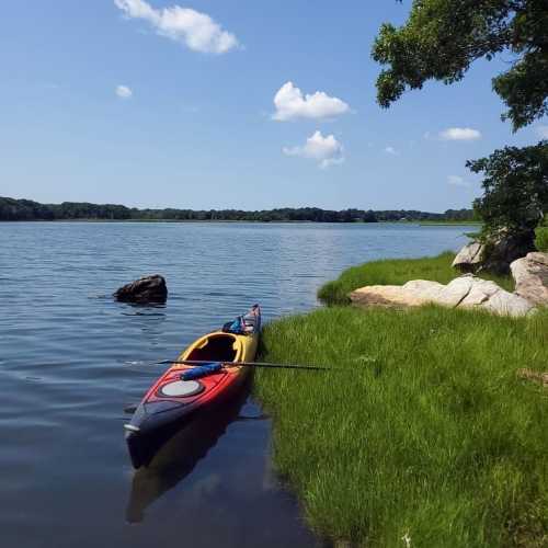 A colorful kayak rests on the shore of a calm lake, surrounded by lush green grass and trees under a blue sky.