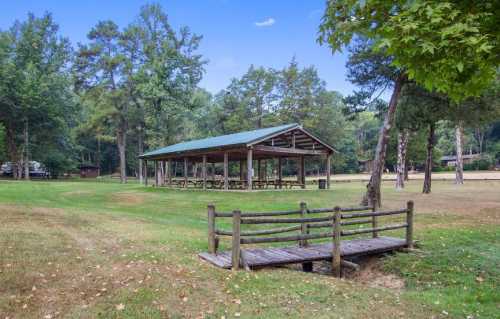 A wooden pavilion with picnic tables surrounded by trees, with a small bridge over a creek in a grassy area.