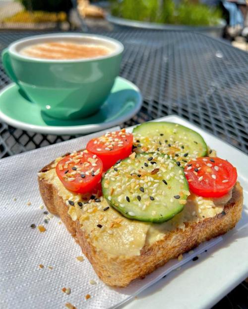 A slice of toast topped with hummus, cucumber, cherry tomatoes, and sesame seeds, with a cup of coffee in the background.