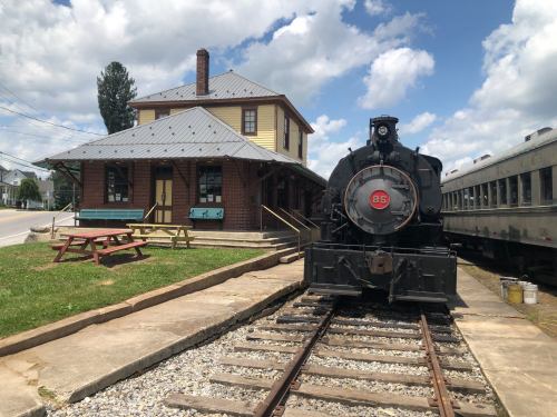 A vintage steam locomotive at a historic train station with a picnic area and blue sky in the background.