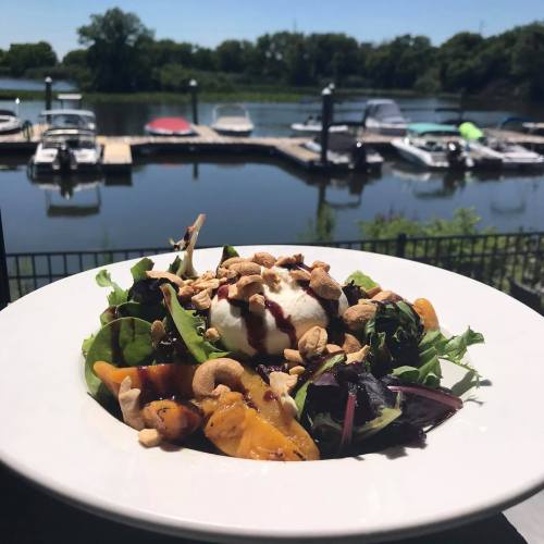 A fresh salad with greens, fruit, and nuts, served in front of a marina with boats on a sunny day.