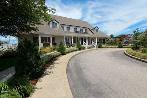 A large, light-colored building with a sloped roof, surrounded by greenery and a curved driveway under a blue sky.