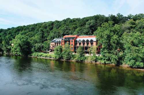 A modern building along a river, surrounded by lush green trees and a hillside under a clear blue sky.