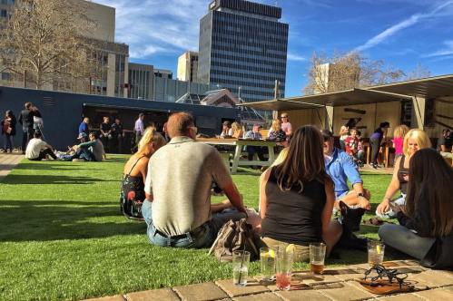 People socializing on a grassy area with drinks, surrounded by buildings and a lively atmosphere.