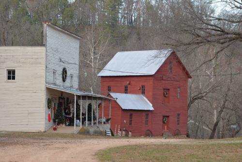 Two rustic buildings, one white and one red, decorated for the holidays, set against a wooded backdrop.