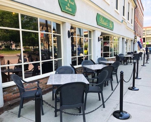 Outdoor seating area of a restaurant with tables and chairs, featuring large windows reflecting the surroundings.
