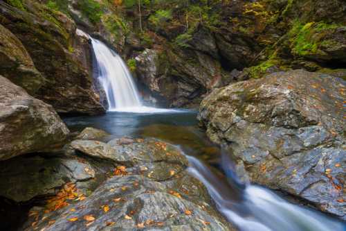 A serene waterfall cascades over rocks into a clear pool, surrounded by autumn foliage and rugged stone formations.