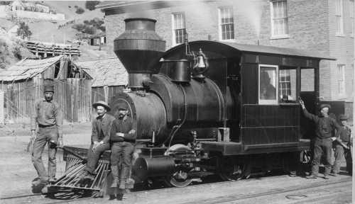 A vintage steam locomotive with five men posing beside it, set against a backdrop of wooden buildings.