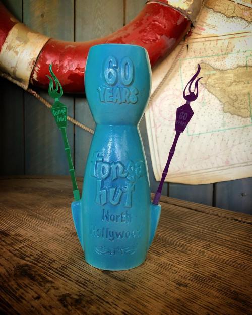 A blue tiki mug celebrating 60 years of Tonga Hut, with colorful torches on either side, set against a rustic background.