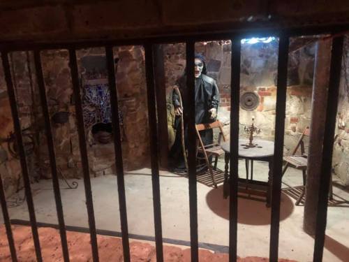 A dimly lit room with a lifelike figure behind bars, surrounded by old furniture and stone walls.