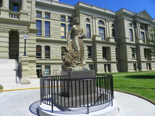 Statue of a woman holding flowers, surrounded by a black fence, in front of a historic stone building.