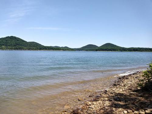 A serene lake view with gentle waves, rocky shore, and green hills in the background under a clear blue sky.