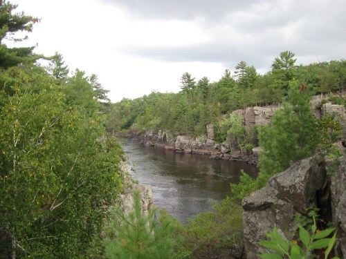 A serene river flows between rocky cliffs, surrounded by lush green trees under a cloudy sky.