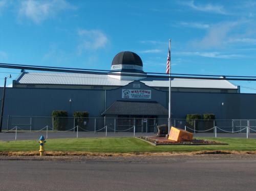 A large gray building with a dome roof, featuring a "Welcome" sign and an American flag in front. Green grass surrounds it.