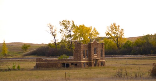 Abandoned stone building in a grassy field, surrounded by trees and distant hills under a pale sky.