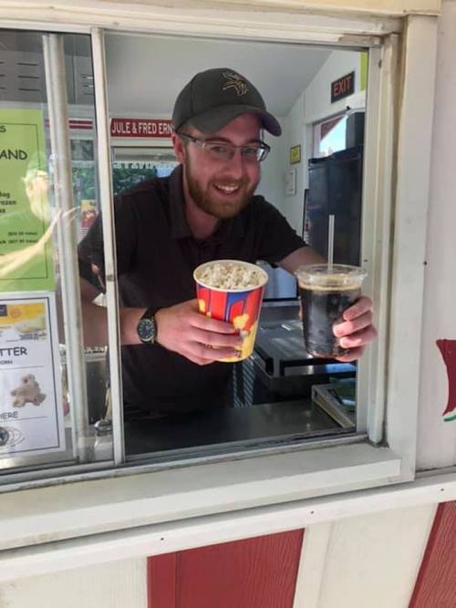 A smiling man at a food stand holds a cup of popcorn and a drink, standing behind a service window.