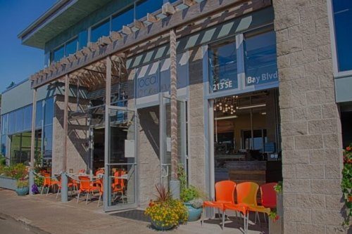 Exterior of a modern restaurant with large windows, orange chairs, and a welcoming entrance on a sunny day.