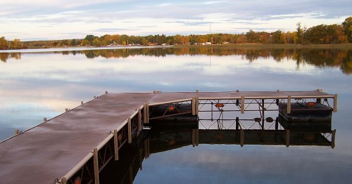 A serene lake scene featuring a wooden dock extending over calm waters, reflecting trees and a cloudy sky.