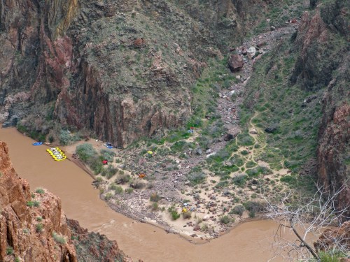Aerial view of a riverbank with rafts, rocky terrain, and greenery along the water's edge in a canyon.