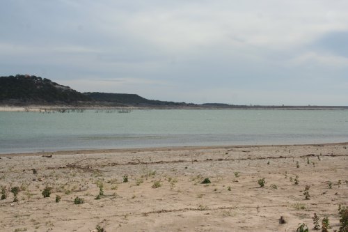 A calm lake with a sandy shore, surrounded by hills under a cloudy sky. Sparse vegetation is visible along the beach.