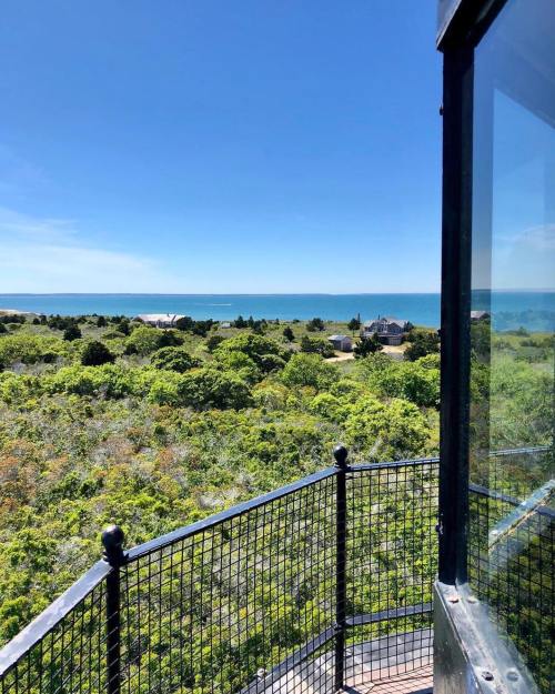View from a lighthouse balcony overlooking lush greenery and a calm blue sea under a clear sky.
