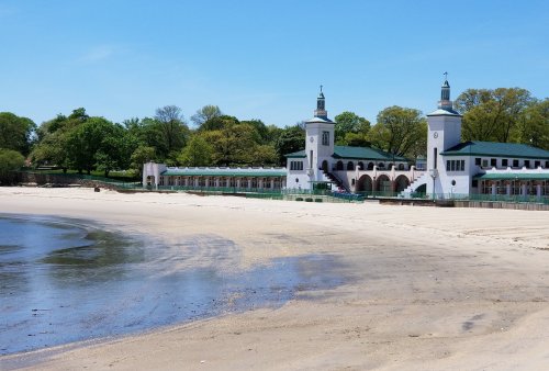 A sandy beach with a calm shoreline and a historic building featuring towers, surrounded by green trees under a clear blue sky.