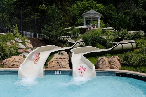 Two children slide down water slides into a pool, surrounded by greenery and a gazebo in the background.
