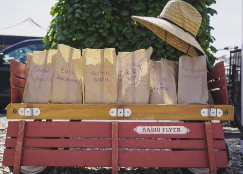 A red wagon filled with paper bags labeled with playful phrases, topped with a straw hat, against a green backdrop.
