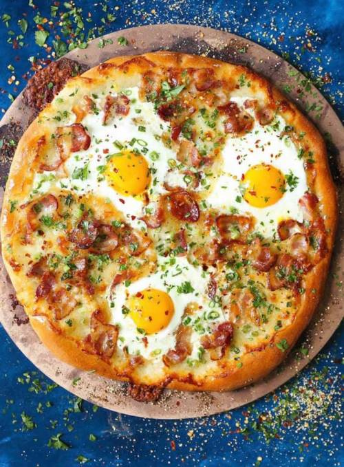 A delicious pizza topped with bacon, herbs, and three sunny-side-up eggs on a wooden board.