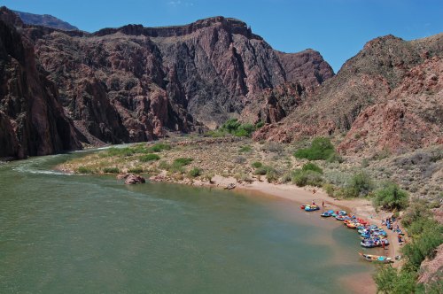 A scenic view of a riverbank with colorful rafts, surrounded by rocky cliffs and lush greenery under a clear blue sky.