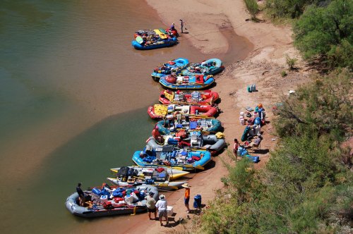 A lineup of colorful rafts on a sandy riverbank, with people preparing for a river adventure.