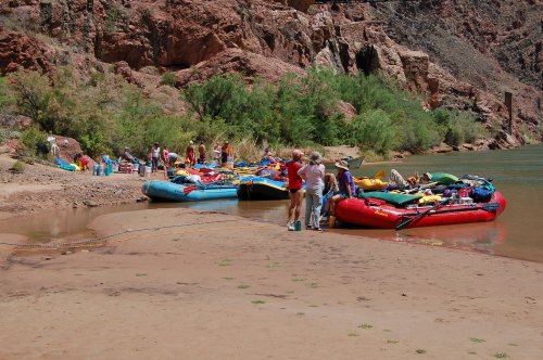 Group of people preparing rafts on a sandy riverbank surrounded by rocky cliffs and greenery.