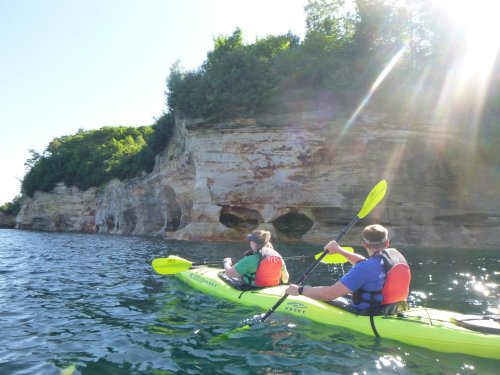Two people kayaking near rocky cliffs under bright sunlight, surrounded by calm water and greenery.