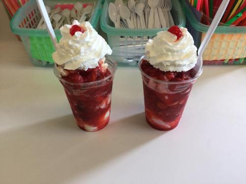 Two cups of strawberry dessert topped with whipped cream and cherries, with colorful utensils in the background.