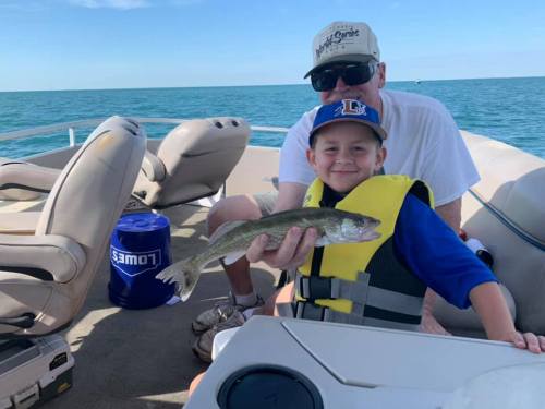 A child in a life jacket proudly holds a fish while sitting on a boat with an adult in a cap.
