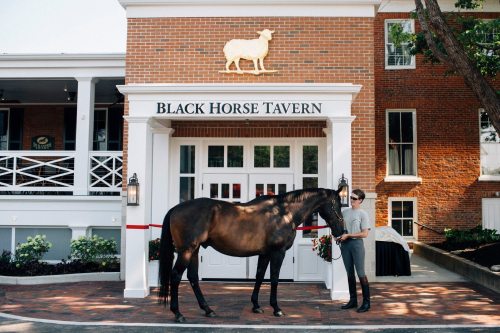A person stands next to a horse in front of the Black Horse Tavern, a brick building with a sheep sign above.