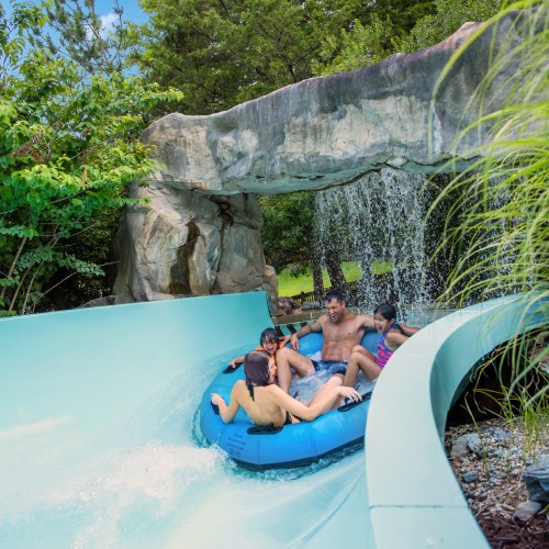 A family enjoys a water slide, splashing through a waterfall in a lush, green setting.
