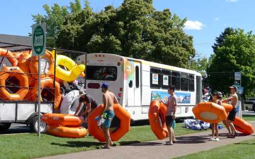 A group of people loading orange inflatable tubes onto a bus near a park on a sunny day.